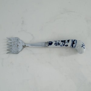 May include: A silver-toned serving fork with a decorative, ornate head and a handle with a blue and white floral design. The handle is made of porcelain. The fork is likely used for serving food.