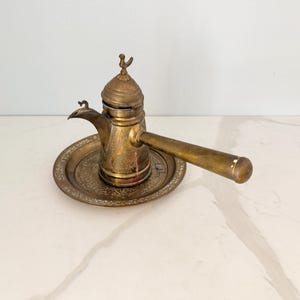 May include: A vintage brass coffee pot with a long handle and a decorative lid, resting on a matching brass plate. The pot has a spout and is adorned with intricate patterns. The overall color is a warm, aged brass.
