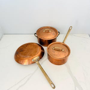 May include: A set of three copper cookware pieces including a frying pan with a long handle, a lidded saucepan with a long handle, and a lidded pot with two side handles. Each piece features polished copper surfaces and brass handles.