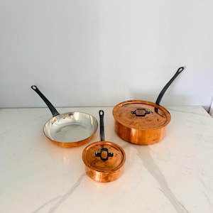 May include: A set of three copper cookware pieces including one open skillet with a silver-toned interior and two lidded pots. Each piece features a polished copper exterior and a long, dark, slender handle attached with metal rivets.