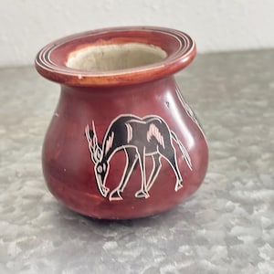 May include: A small, round, maroon-colored vase with a wide rim. The vase features a black and white etched design of an antelope. The interior of the vase is a lighter beige color.