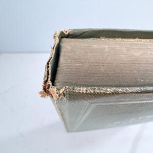 Puede incluir: Primer plano de la esquina de un libro antiguo, mostrando la cubierta y las p&aacute;ginas desgastadas. El libro tiene una cubierta verde descolorida con desgaste visible en los bordes, lo que sugiere su edad y uso.