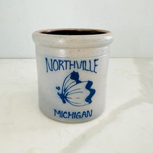 Op de afbeelding: Een cilindrische steengoed pot met een lichtgrijze buitenkant en een donkerbruine binnenkant. De pot heeft blauwe letters met de tekst "NORTHVILLE MICHIGAN" en een blauw vlinderontwerp.