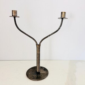 May include: A dual-arm metal candelabra featuring a circular base, a central stem wrapped in fine wire, and two curved arms extending upward to support candle cups. The metal surface displays a dark, weathered patina with visible signs of oxidation.
