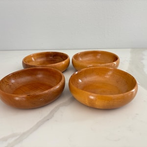 May include: Four small, oval-shaped wooden bowls with a warm, light brown color. The bowls are arranged in a square formation, each with a smooth, polished surface. The wooden bowls are likely for serving snacks or small portions.