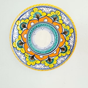 May include: A round ceramic plate featuring a vibrant hand-painted design. The center has a teal and white circular pattern, surrounded by a ring of bright yellow and orange floral motifs with black line details. The outer rim is yellow.