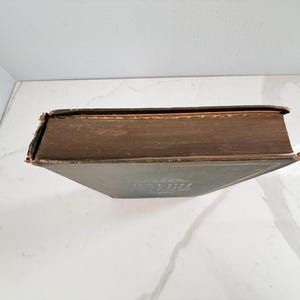 Puede incluir: Un libro antiguo con una cubierta verde oscuro y p&aacute;ginas envejecidas con tonos marrones. El lomo y los bordes del libro muestran signos de desgaste, lo que sugiere su edad. El t&iacute;tulo es visible en la portada. El libro est&aacute; sobre una superficie blanca.