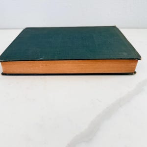 Puede incluir: Un libro antiguo de tapa dura con una cubierta verde oscuro y p&aacute;ginas de color naranja. El libro est&aacute; cerrado y descansa sobre una superficie blanca. Los bordes de las p&aacute;ginas son visibles y el lomo no es visible.