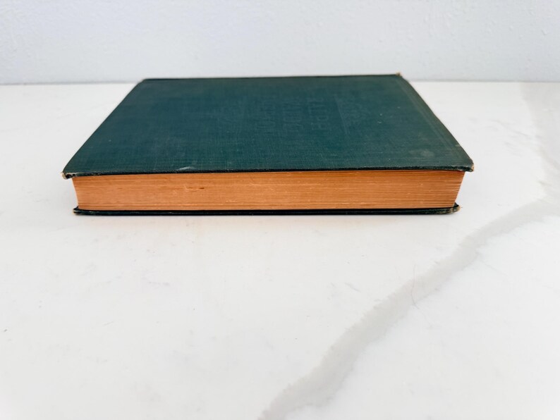 Puede incluir: Un libro antiguo de tapa dura con una cubierta verde oscuro y p&aacute;ginas de color naranja. El libro est&aacute; cerrado y descansa sobre una superficie blanca. Los bordes de las p&aacute;ginas son visibles y el lomo no es visible.