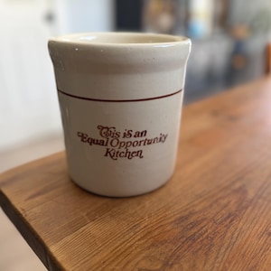 May include: A cream-colored ceramic crock featuring a thin brown horizontal line near the top rim and the brown text "This is an Equal Opportunity Kitchen" printed on the front. The vessel has a cylindrical shape with a slightly flared rim.