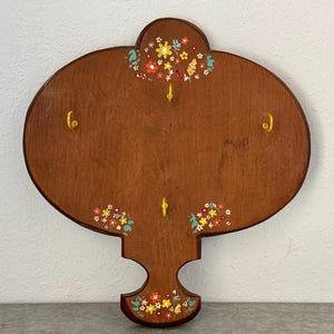 May include: A wooden wall-mounted coat rack with a unique, decorative shape. The brown rack features four gold-colored hooks and is adorned with colorful floral accents in yellow, red, and blue. The rack is designed to hold coats, hats, or other items.