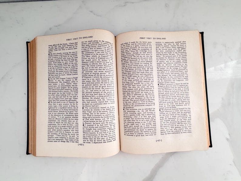 Puede incluir: Un libro abierto con el t&iacute;tulo "FIRST VISIT TO ENGLAND" impreso en la parte superior de cada p&aacute;gina. Las p&aacute;ginas est&aacute;n llenas de texto negro peque&ntilde;o. El libro tiene una cubierta negra y est&aacute; sobre un fondo blanco.