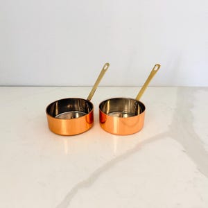 May include: Two small, round saucepans with polished copper exteriors and silver-toned metallic interiors. Each pan features a single, long, slender brass-colored handle extending upward at an angle from the side of the circular vessel.