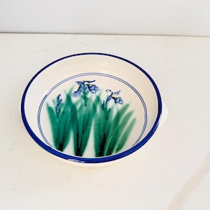 May include: A round ceramic bowl with a cream-colored base and a dark blue rim. The interior features a hand-painted design of green grass blades and blue iris flowers. A thin blue circular line accents the inner rim of the bowl.