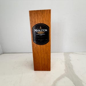 May include: A wooden box with a dark label that reads "Midleton Very Rare Irish Whiskey". The box is rectangular and made of light brown wood. The label is black with gold lettering and a gold border. The box is standing upright.