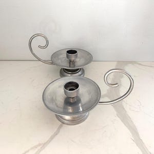 May include: Two silver candle holders with a classic design. Each holder features a circular base, a central cup for the candle, and a curved handle. The candle holders are made of a shiny, metallic material and are placed on a white surface.