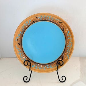 May include: A decorative ceramic plate with a light blue center and a brown rim. The rim features a painted design with blue, white, and black accents. The plate is displayed on a black metal stand. The plate is a decorative item.