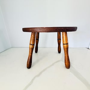 May include: A small, round wooden stool with a dark brown, circular top and four turned, light brown legs. The stool features a visible wood grain texture throughout, with a smooth, flat surface and tapered legs that provide a stable base.