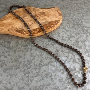 Puede incluir: Un collar de cuentas marrón con un cierre dorado. El collar está hecho de pequeñas cuentas facetadas y se muestra sobre una superficie de madera. Las cuentas son de un color oscuro y terroso, y el collar está dispuesto en forma curva.