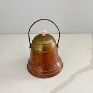 May include: A vintage copper and brass bell-shaped container with a handle. The top is brass, and the body is copper with horizontal ridges. It has a small, off-white knob on top. The handle is a curved brass wire.