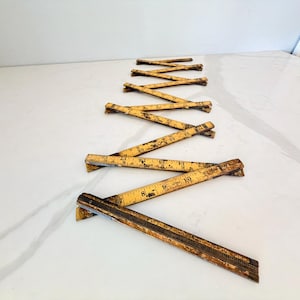 May include: A vintage yellow folding wooden ruler with black measurement markings in inches. The ruler is partially extended in a zigzag pattern, showing worn paint and metal hinges connecting the individual segments of the measuring tool.