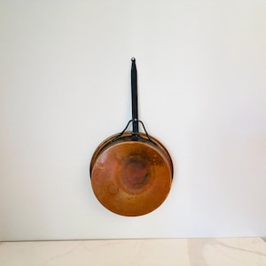 May include: A round copper pan with a weathered, metallic orange-brown finish. The pan features a long, slender black metal handle attached with a Y-shaped support bracket, extending vertically from the top edge of the circular cooking vessel.