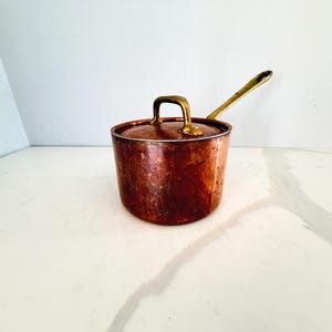 May include: A copper saucepan with a lid and a long handle. The pot has a hammered texture and a brass handle and lid handle. The handle is also brass. The pot is sitting on a white surface.