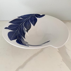 May include: White ceramic bowl with a navy blue leaf design. The bowl has a wide, shallow shape with a slightly curved edge. The leaf pattern is on the left side of the bowl. The bowl is sitting on a white surface.
