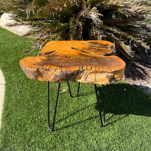 May include: A side table featuring a natural live edge wood slab top with a glossy amber finish, prominent dark wood grain, and deep cracks. The tabletop is supported by four thin, black metal hairpin legs that provide a stable base for the piece.