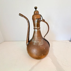 May include: A copper pitcher with a rounded base, a long slender neck, and a curved spout. The metal surface shows signs of oxidation and wear, featuring a matching lid with a small knob handle and a thin, curved handle attached to the side.