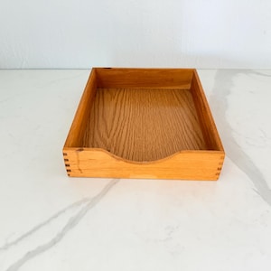May include: A rectangular wooden tray with a natural wood grain finish. The tray has a cutout on one side and is constructed with visible dovetail joints. The tray is empty and sits on a white marble surface.