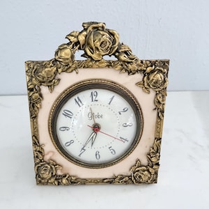 May include: A vintage decorative clock with a square face and a cream-colored background. The clock face has black numerals and hands, with a red second hand. The clock is framed with gold-colored roses.