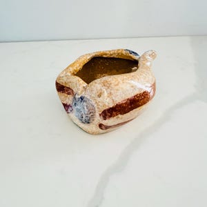 May include: A small, irregularly shaped ceramic bowl with a glossy, speckled beige finish. The exterior features horizontal brown stripes and a circular blue accent. The interior is a smooth, dark brown, and the rim has a wavy, organic edge.