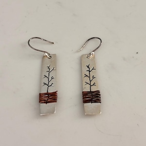 May include: A pair of silver-tone earrings. Each earring features a rectangular silver plate with a tree design and wrapped copper wire. The earrings have a hook closure. The background is a light, neutral color.
