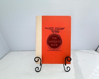 May include: A vintage book titled "The Antiques Book" with a red cover and an eagle emblem. The book is displayed on a black metal stand with scrolled details. The spine is a light beige color.