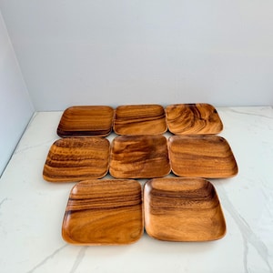 以下が含まれることがあります： 角が丸い正方形の木製プレート8枚のコレクション。 プレートは明るい茶色の木材でできており、顕著な木目模様があります。 白い表面に配置されています。