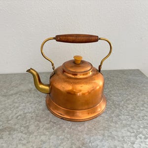 Puede incluir: Una tetera de cobre con asa de madera y detalles en latón. La tetera tiene un cuerpo redondeado, un pico y una tapa con un pequeño pomo. El asa es arqueada y la tetera se asienta sobre una base plana.