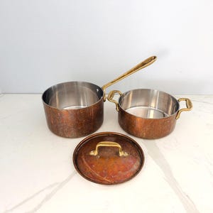 May include: A copper-colored saucepan with a long brass handle, a copper-colored pot with two brass handles, and a copper-colored lid with a brass handle. The interiors of the pots are stainless steel. The cookware is on a white surface.