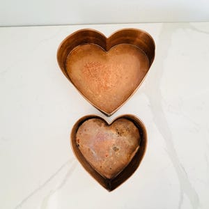 May include: Two heart-shaped copper baking pans of different sizes. The larger pan is positioned above the smaller pan. Both pans feature a metallic copper finish with visible signs of wear, oxidation, and discoloration on the interior surfaces.