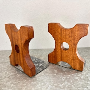 May include: Pair of wooden bookends. Each bookend is a warm brown color with a unique shape, featuring a central circular cutout and a metal base. The bookends are designed to hold books upright on a shelf or desk.