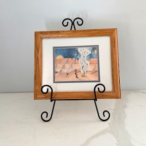 May include: A framed watercolor painting depicting a desert scene with adobe structures, a figure, and rising smoke. The artwork is set in a light brown wooden frame and displayed on a black metal easel with decorative scrollwork.
