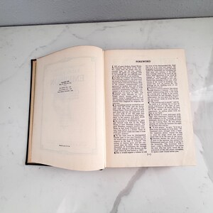 Puede incluir: Un libro abierto con lomo negro y p&aacute;ginas blanquecinas. La p&aacute;gina izquierda tiene un borde decorativo y texto. La p&aacute;gina derecha presenta la palabra "FOREWORD" en la parte superior y columnas de texto. El libro est&aacute; sobre una superficie blanca.