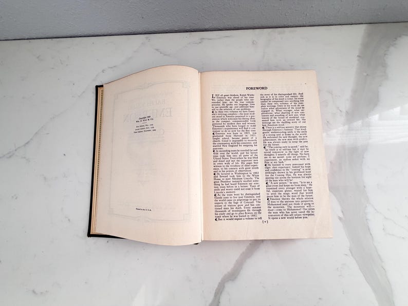 Puede incluir: Un libro abierto con lomo negro y p&aacute;ginas blanquecinas. La p&aacute;gina izquierda tiene un borde decorativo y texto. La p&aacute;gina derecha presenta la palabra "FOREWORD" en la parte superior y columnas de texto. El libro est&aacute; sobre una superficie blanca.