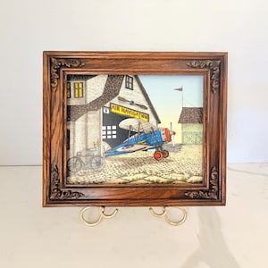 May include: A framed illustration featuring a blue biplane parked in front of a building labeled AIR NAVIGATION. The scene includes a bicycle, a cobblestone ground, and a small outbuilding. The artwork is enclosed in a dark brown ornate wooden frame.