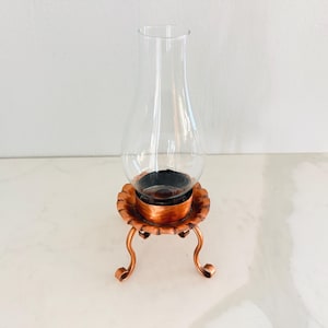May include: A vintage copper-toned oil lamp with a clear glass chimney. The lamp features a decorative base with a scalloped edge and three curved legs. The lamp is designed to hold oil for illumination.