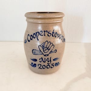 May include: A tan stoneware jar with a rounded body and a slightly flared rim. The jar is decorated with blue lettering that reads "Cooperstown NY 2003" and a baseball glove, bat, and ball design. The jar has a rustic, vintage aesthetic.