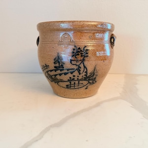 May include: A tan stoneware crock with a dark blue floral design. The antique pot has two small handles and a decorative band near the top. The crock is a round shape and sits on a white surface.
