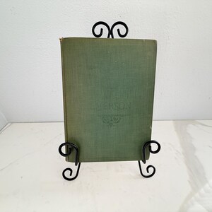 Puede incluir: Un libro antiguo con una cubierta verde se exhibe en un soporte de metal negro. La cubierta del libro tiene texto en relieve y un dise&ntilde;o decorativo. El soporte tiene un dise&ntilde;o en espiral. El libro es un art&iacute;culo decorativo cl&aacute;sico.