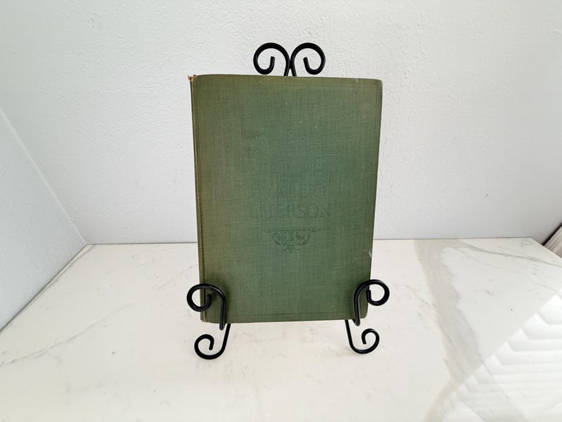 Puede incluir: Un libro antiguo con una cubierta verde se exhibe en un soporte de metal negro. La cubierta del libro tiene texto en relieve y un dise&ntilde;o decorativo. El soporte tiene un dise&ntilde;o en espiral. El libro es un art&iacute;culo decorativo cl&aacute;sico.