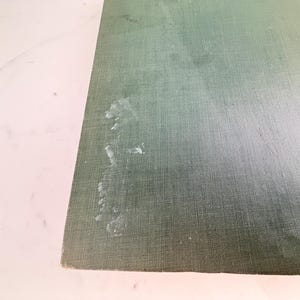 Puede incluir: Primer plano de una secci&oacute;n de la cubierta de un libro. La cubierta es de color verde apagado con un aspecto de tela texturizada. Hay varias manchas blancas en la cubierta. El libro est&aacute; sobre una superficie blanca.